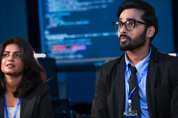 An Indian developer holds a tablet while a Caucasian engineer interacts with the screen. They are analyzing AI data or a cybersecurity system in a SOC.