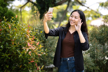 Asian woman waving on smartphone video call in garden, remote communication