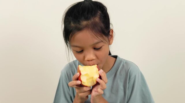 A video shows a girl standing holding a red apple and biting it bit by bit, almost halfway through. With different poses and perspectives, she smiles brightly as she enjoys eating her favourite fruit.