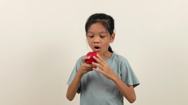 A video shows a girl standing holding a red apple and biting it bit by bit, almost halfway through. With different poses and perspectives, she smiles brightly as she enjoys eating her favourite fruit.