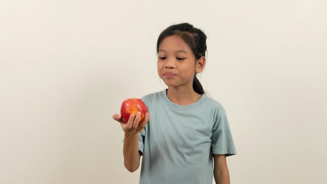 A video shows a girl standing holding a red apple and biting it bit by bit, almost halfway through. With different poses and perspectives, she smiles brightly as she enjoys eating her favourite fruit.