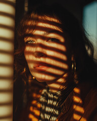 Woman with thoughtful expression is illuminated by sunlight filtered through blinds, creating striped shadows on her face. She wears hoop earrings and patterned top, adding to artistic composition