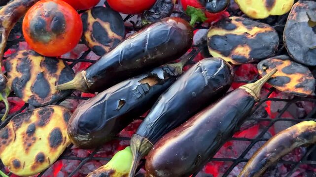A close-up of whole eggplants, sliced vegetables, and tomatoes developing charred marks on a hot grill grate, creating an appetizing texture for smoky, rustic cooking.