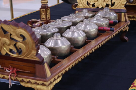 A perspective close-up of a Bonang Barung or Panerus, a Javanese or Balinese gamelan instrument with a row of small, highly polished bronze gongs on a carved wooden frame.