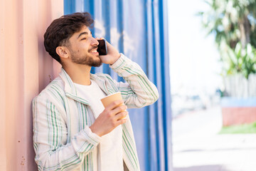 Handsome Arab man at outdoors using mobile phone and holding a coffee with happy expression