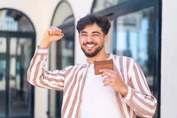 Handsome Arab man at outdoors holding a wallet and celebrating a victory