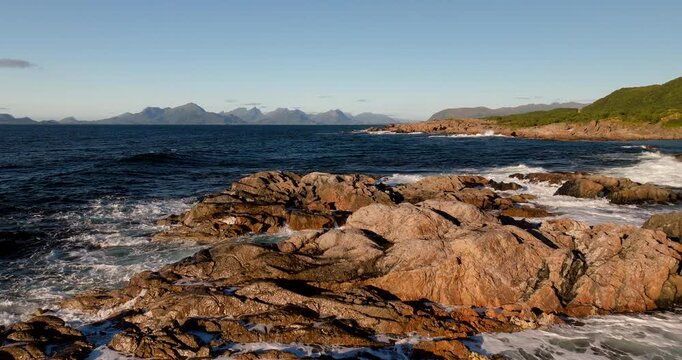 Aerial view of crashing waves over rocky coastline near Uv&aelig;rshula and Stokmarknes in northern Norway. Dramatic arctic shoreline with ocean spray, rugged terrain, and scenic coastal mountains