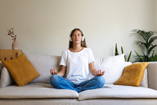 Front view of young woman meditating on the couch listening to guided meditation using wireless headphones.