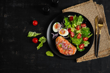 Delicious healthy breakfast - boiled egg, sandwich salmon and green salad. Balanced meal plate on dark background, top view, copy space.