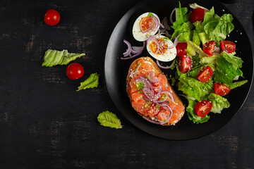Delicious healthy breakfast - boiled egg, sandwich salmon and green salad. Balanced meal plate on dark background, top view, copy space.