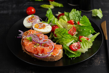 Delicious healthy breakfast - boiled egg, sandwich salmon and green salad. Balanced meal plate on dark background