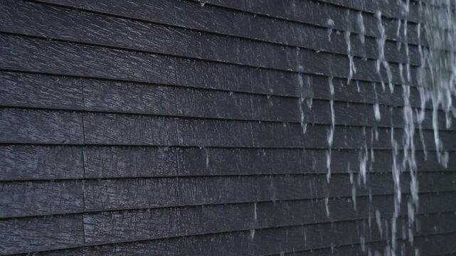 Melancholic rain water running down dark grey wall siding during gloomy storm. This dramatic, wet downpour cascades over textured stone, creating somber outdoor scene