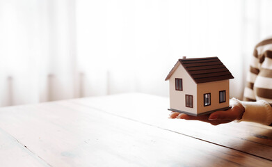 A person holding a small model house on their hand above a wooden table, symbolizing real estate, home ownership, and property investment.