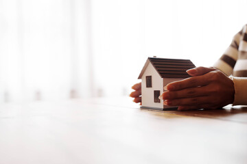 A person holding a small model house on their hand above a wooden table, symbolizing real estate, home ownership, and property investment.