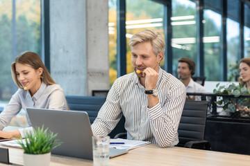Two call center employees, woman and a man, are working on laptops at their desks while wearing headsets, with colleagues in the background.