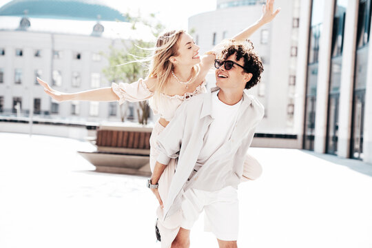 Young smiling beautiful woman and her handsome boyfriend in casual summer clothes. Happy cheerful family. Couple posing in street at sunny day. Female sits at his back. piggyback riding, raising hands