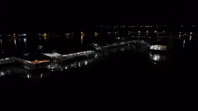 Aerial b roll night scene of buildings and pier over dark water with scattered lights reflecting on the surface creating a calm moody and cinematic nighttime atmosphere