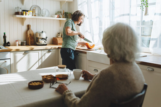 Rare view of senior lady sitting at kitchen table, having herbal tea looking at nurse or caregiver cutting pumpkin standing next to counter, having nice conversation while social worker cooking