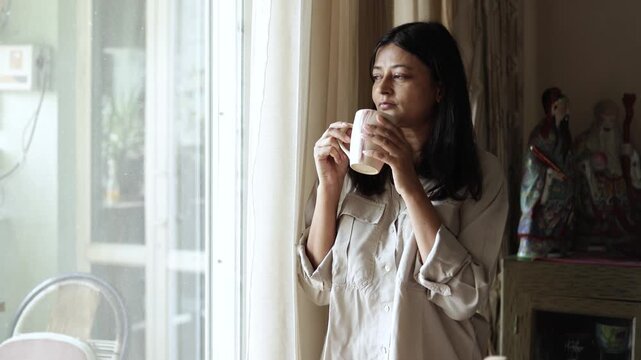 Young indian woman enjoying morning tea or coffee looking out of windrow at home. Daily routine. 4k