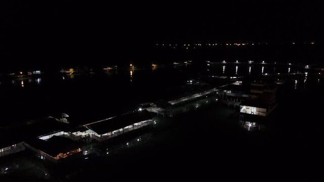 Aerial b roll night scene of buildings and pier over dark water with scattered lights reflecting on the surface creating a calm moody and cinematic nighttime atmosphere