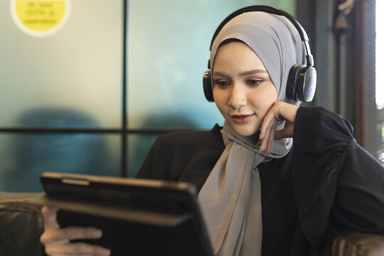Muslim woman wearing headphones and hijab watching content on digital tablet. Concept of online learning, remote work, streaming media, digital lifestyle and modern education technology.
