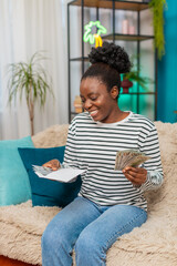 African American Black woman counting money bills at home. Sitting on sofa, girl looks satisfied,...