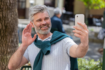 Bearded mature man blogger using smartphone, communicating video call online with subscribers, recording stories for social media vlog outdoors. Guy sitting on chair in urban sunshine city street.