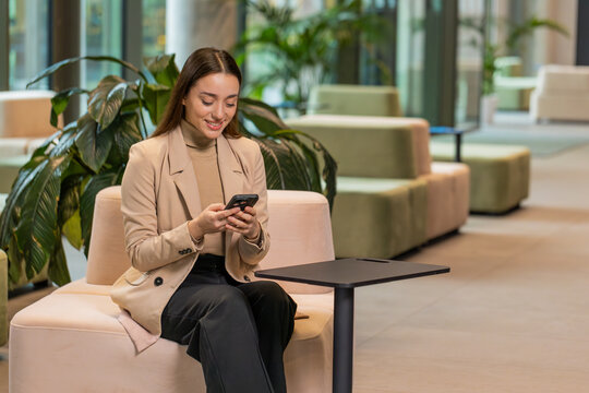 Caucasian young businesswoman girl using mobile phone texting share messages content working on smartphone social media applications online, relaxing in modern office lobby. Lady sitting in hotel hall - Powered by Adobe