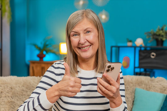 Caucasian mature woman smiles, engages with social media photos videos on smartphone while sitting at home. Grandmother gives thumbs up gesture expressing approval and happiness on sofa in living room - Powered by Adobe