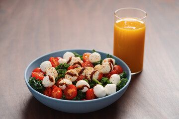 Blue bowl of salad with mozzarella, cherry tomatoes and kale, seasoned with balsamic sauce on walnut wood table with glass of orange juice