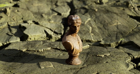 A small antique bust sculpture stands alone amidst cracked earth, revealing a stark contrast of detail against a rough surface. The bright daylight highlights its features and setting.