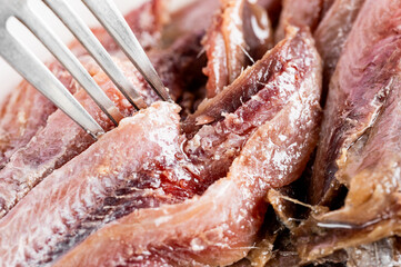 A close-up shot of an oily, salty anchovy fillet being lifted with a fork. The fish is ready to be eaten, suggesting a gourmet or Mediterranean dish