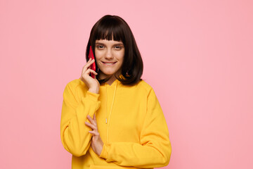 Portrait of a cheerful person wearing a bright yellow hoodie, speaking on a mobile phone against a pastel pink backdrop, conveying casual style, friendly vibe, and modern everyday life.
