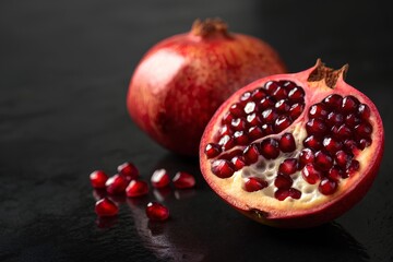 Whole and cut pomegranates with scattered seeds on dark reflective surface, highlighting vibrant red arils and rich natural textures