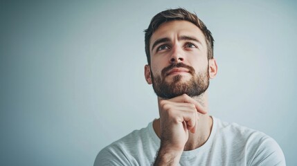 A man with a beard and mustache, wearing a white t-shirt, looking up thoughtfully against a light blue background.