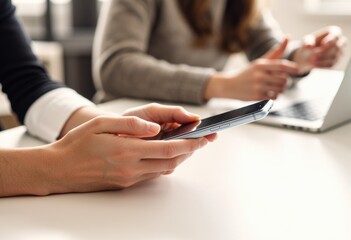 Hands holding a smartphone while another person uses a laptop nearby