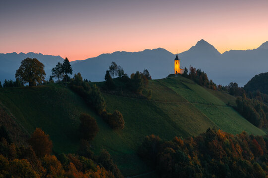 Scenic sunrise view of the Church of St. Primus and Felician in Jamnik, Slovenia. The iconic hilltop church is illuminated by warm morning light, surrounded by misty valleys and the majestic Julian Al