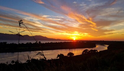 Sunset over a river, with vibrant orange and blue sky, grass silhouetted in the foreground, calm water reflecting light