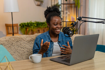 African American young blogger woman sitting at table on home sofa records a personal podcast using a studio microphone, gesturing confidently. Black girl smiles warmly and enjoys sharing voice online