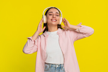 Young woman dancing with closed eyes while listening to music in headphones showing carefree and joyful emotion. Girl isolated on yellow background expresses freedom, relaxation positive peaceful mood