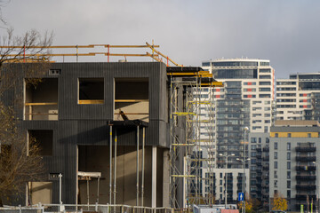 Modern residential building under construction with scaffolding and concrete structure in front of high-rise apartment towers showing urban development progress