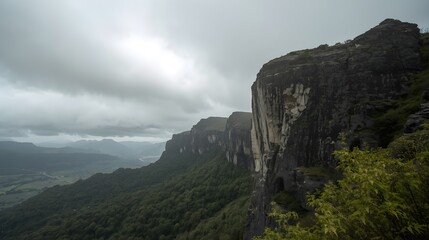 Naklejka premium Dramatic dark rocky mountain cliff face with lush green vegetation under a moody overcast sky with shafts of sunlight breaking through the clouds