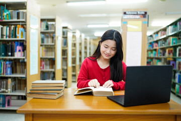 Teenage asian female pretty student whit laptop, studying while sits at the table in a college library, reads books to searching information for a lesson or exam, doing homework