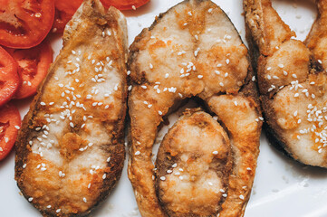 Pieces of sesame-fried fish served on a white plate with sliced tomato. A perfect dish for breakfast, lunch, or dinner.