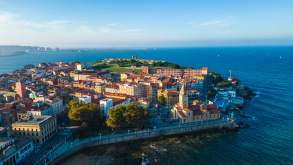 An aerial view captures the vibrant coastal city of Gijon, Spain, showcasing its historic architecture, bustling promenade, and the deep blue waters of the Cantabrian Sea.