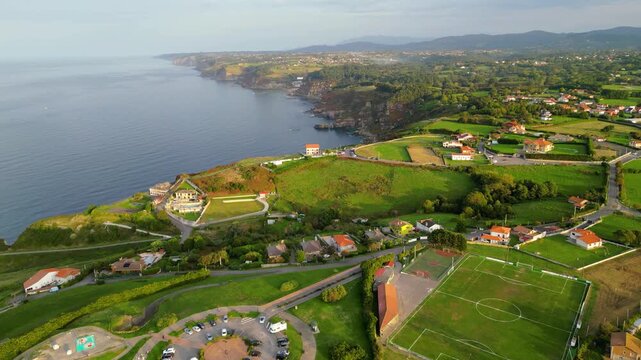Aerial view of the stunning Asturian coastline at golden hour, featuring lush green fields, charming houses, and the tranquil Bay of Gij?n under a warm sunset glow.