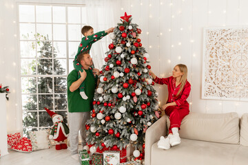 Happy family decorating Christmas tree in living room at home. A little boy sits on dad's shoulders...