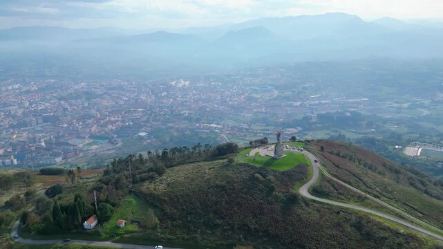 Panoramic aerial view of Oviedo city and the surrounding Asturian landscape, featuring the iconic Cristo del Monte Naranco statue under a cloudy autumn sky.