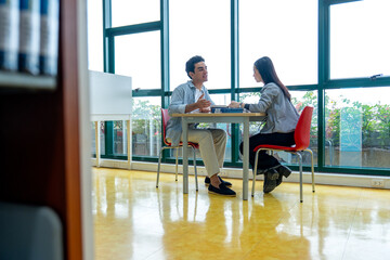 Side view and wide shot Hispanic man sit and discuss with Asian woman at table in public library in front of glass window with day light.