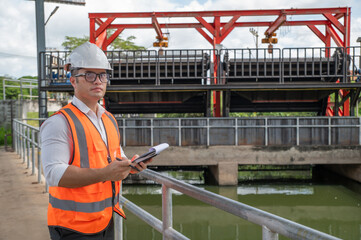 an engineer in safety vest inspects water gate structure, recording data on a sunny day